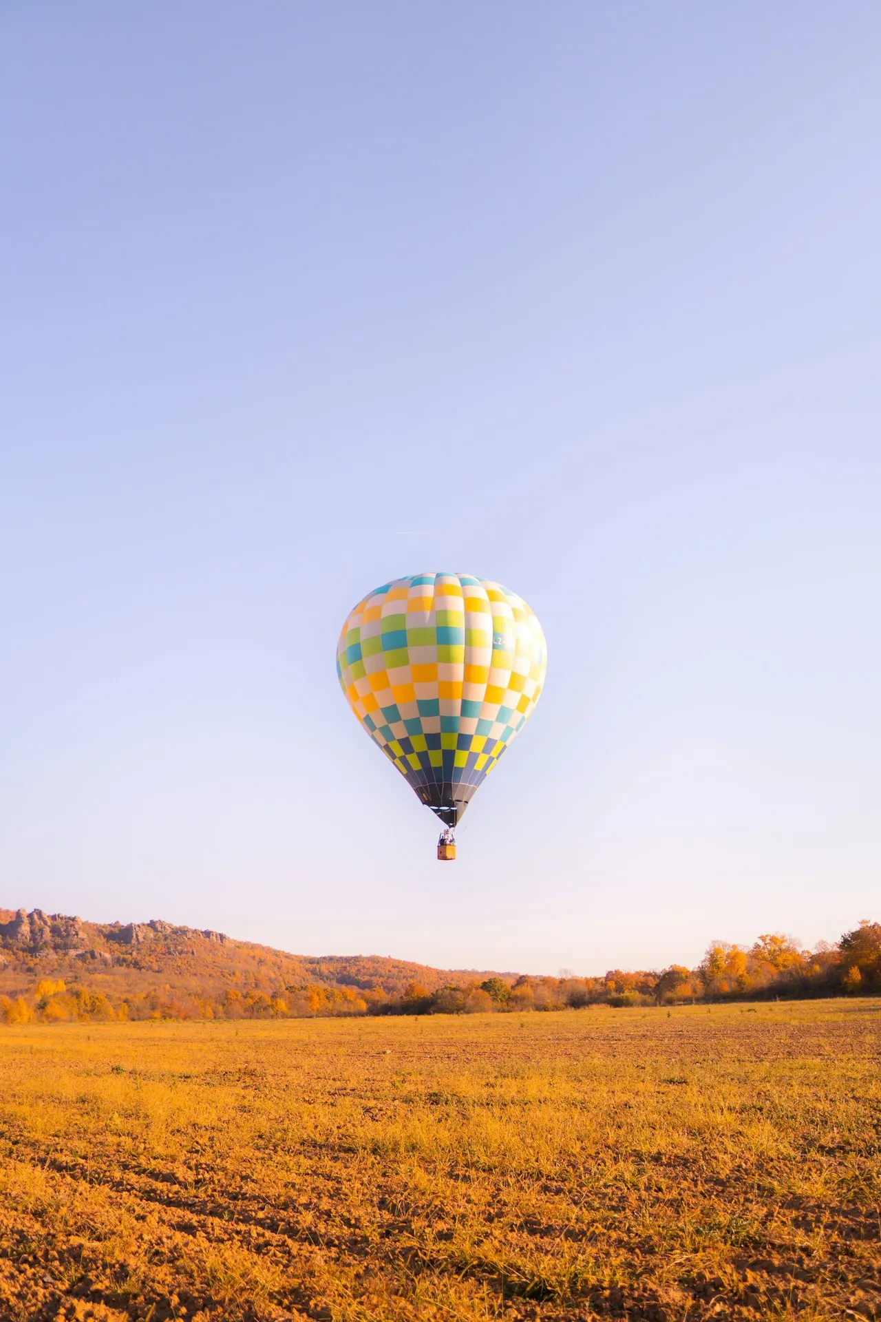 Yellow and teal checkered hot air balloon hovering low over golden autumn farmland with wooded hills in the background