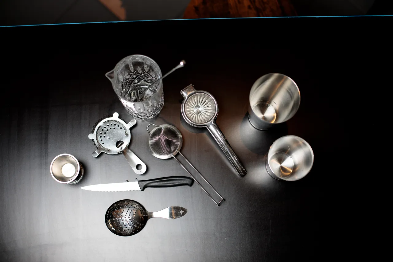 Overhead flat-lay of bartending tools, including strainers, jiggers, a citrus juicer, a knife, and a mixing glass with a barspoon.