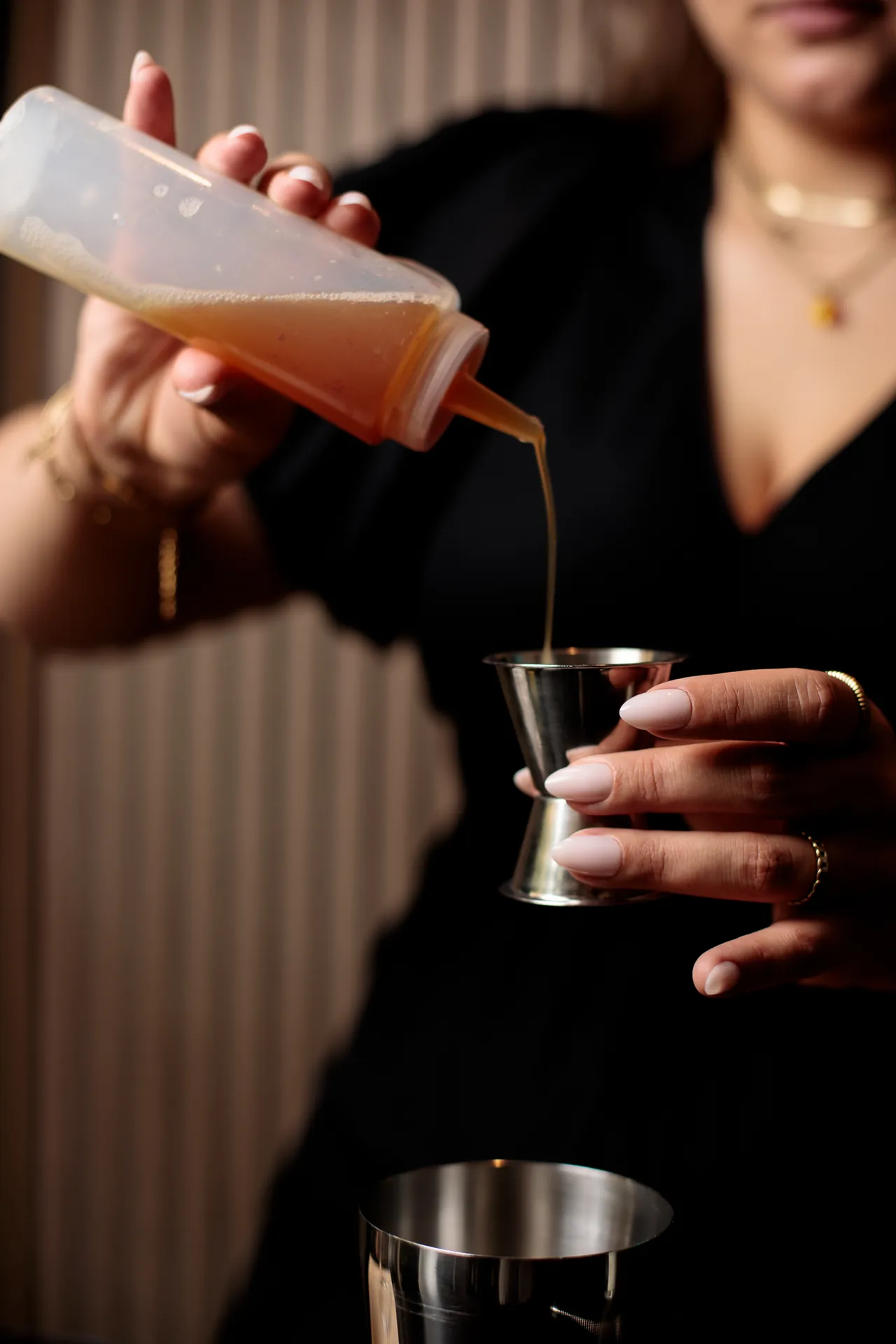 Close-up of hands pouring an orange-hued liquid from a bottle into a jigger to measure a cocktail ingredient.