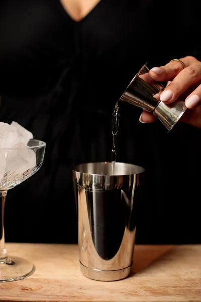 Close-up of hands pouring a measured spirit from a jigger into a cocktail shaker, next to a bowl of ice.