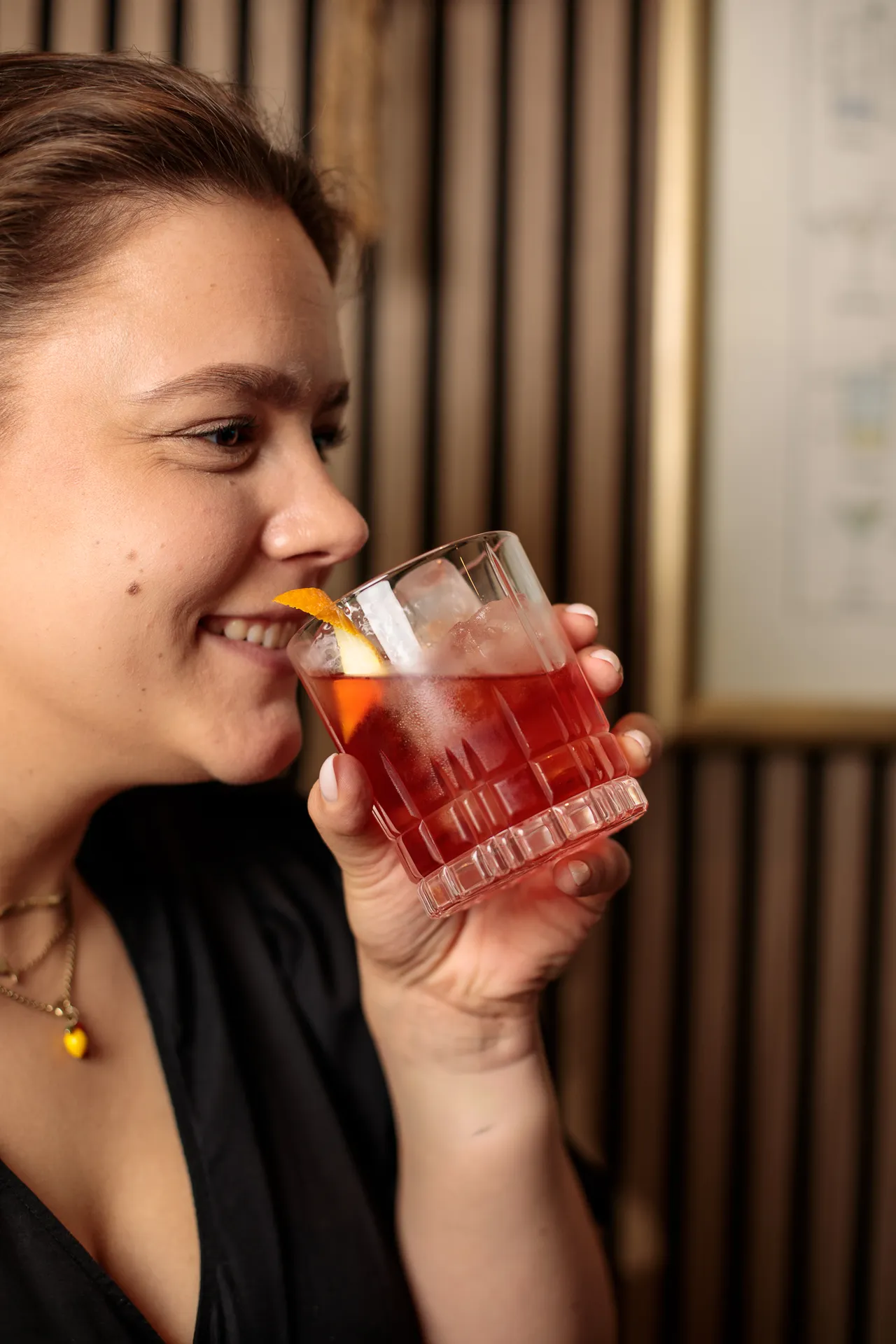 Close-up of a smiling woman sipping a red cocktail garnished with an orange peel.