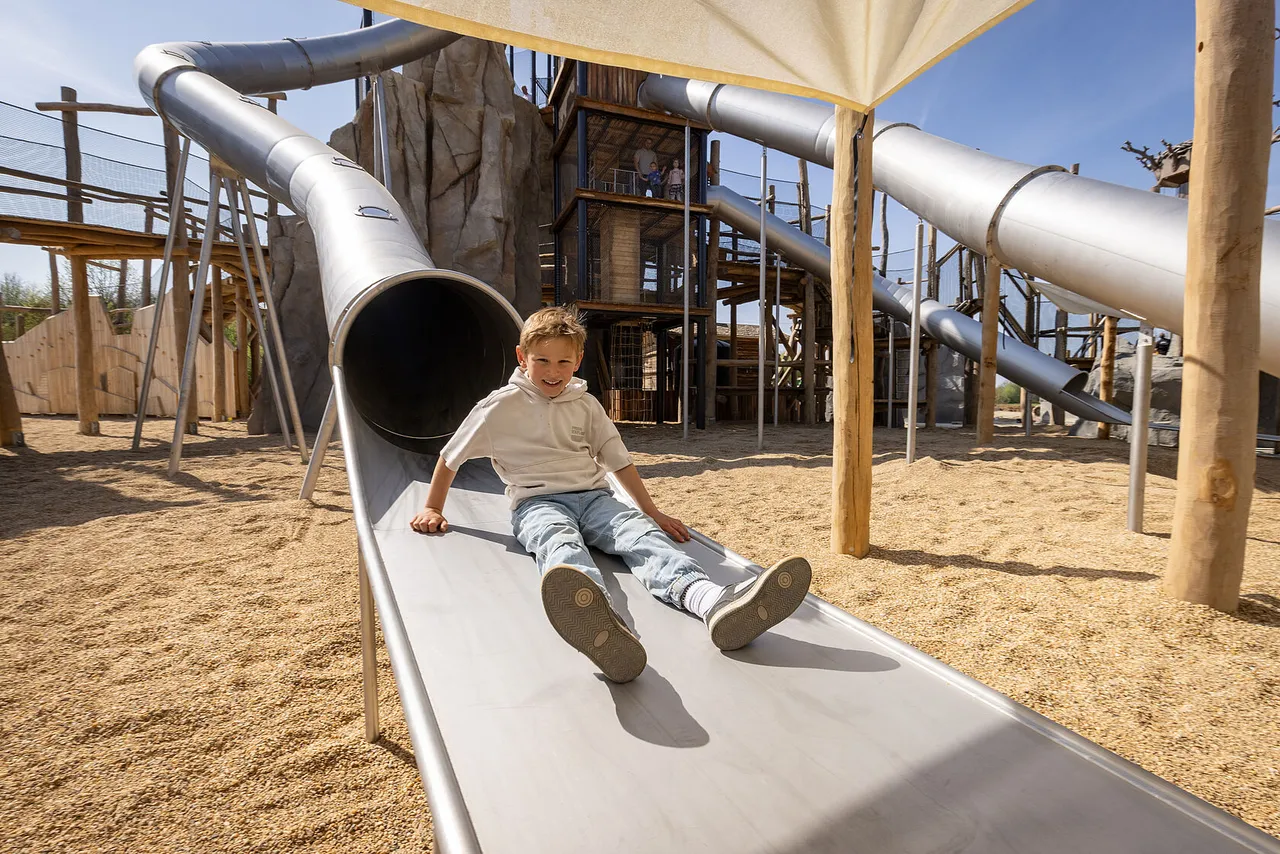 Smiling boy sliding down a wide metal slide at an adventure playground