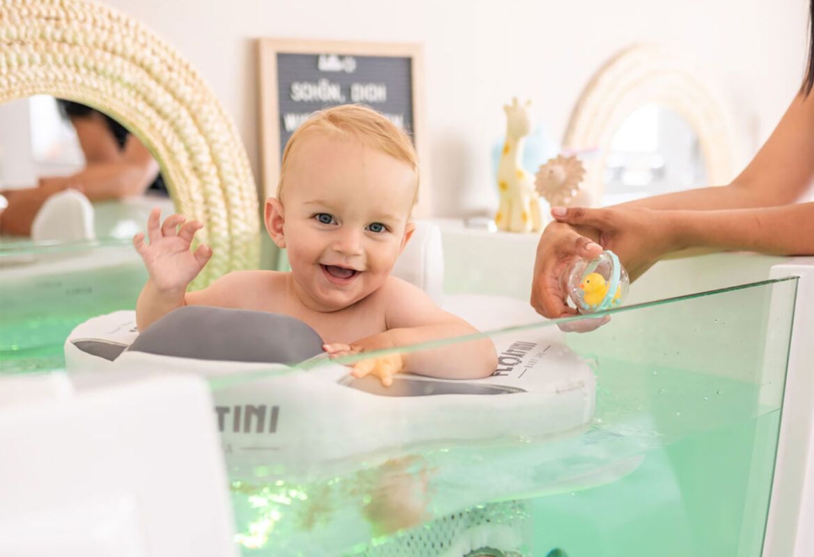 Baby smiling and waving while floating in a baby spa ring, reaching for a rubber duck toy
