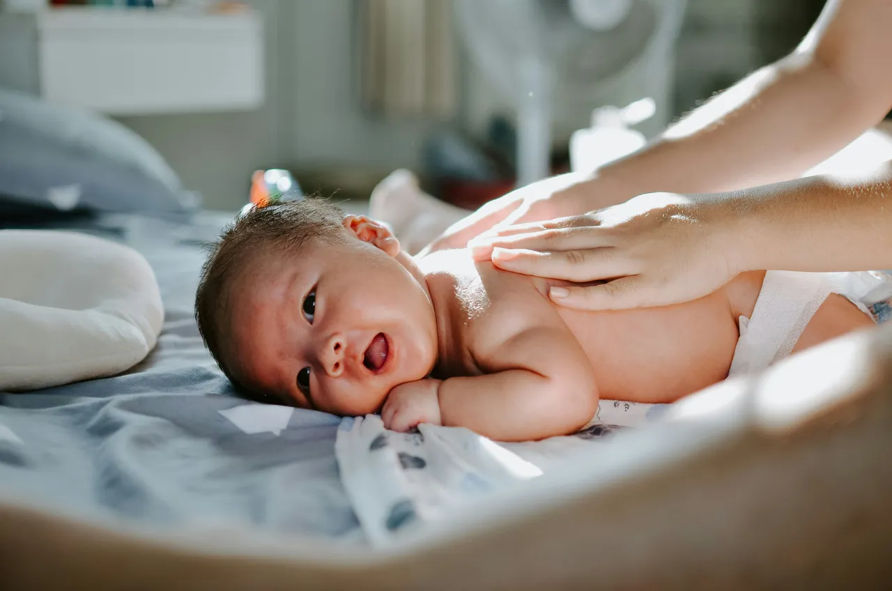 Newborn baby lying on a changing table smiling while a caregiver's hands gently massage its back.