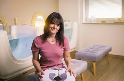 Woman in a pink t-shirt smiling while seated between two baby spa tubs, holding a lavender flotation ring.