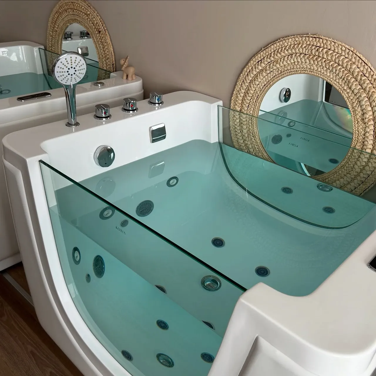 Close-up of a baby spa tub filled with clear blue-green water, showing jet nozzles, a chrome shower handle, and a woven mirror frame.