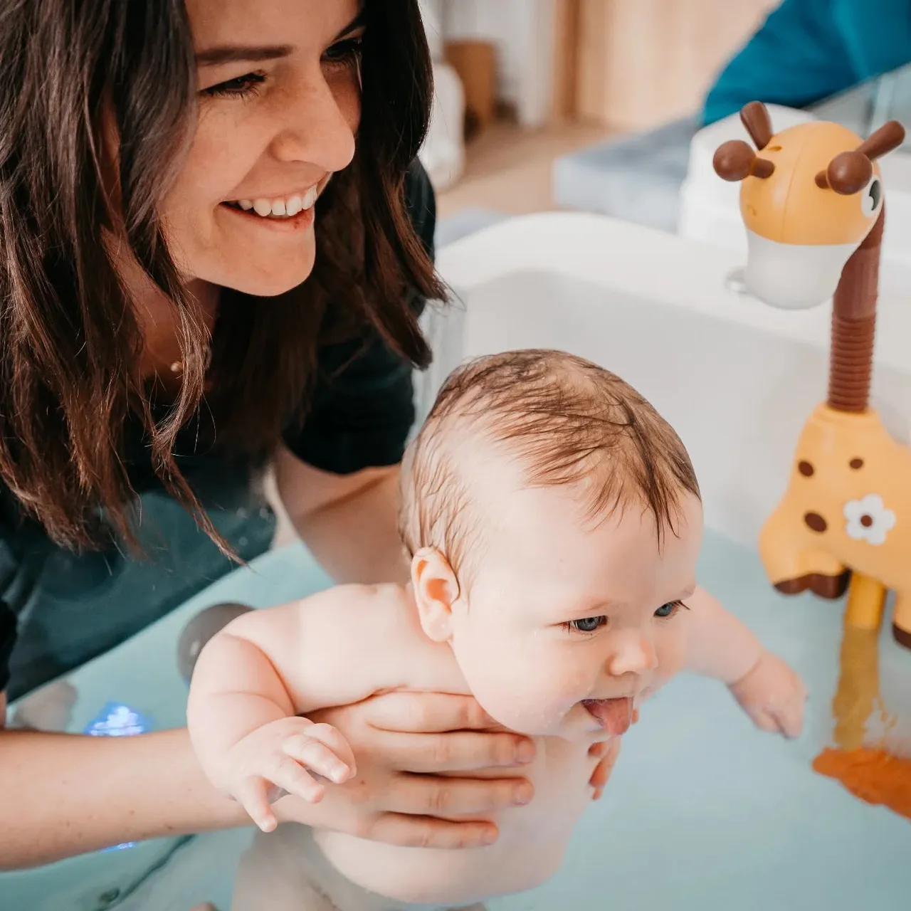 Woman smiling while holding a baby in a baby spa tub, the baby sticking out its tongue, with a giraffe toy nearby.