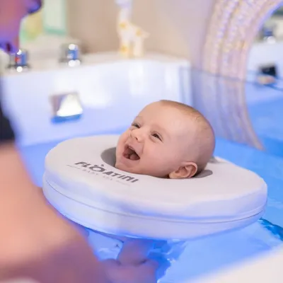 Close-up of a smiling baby's face resting in a white Floatini flotation ring, floating in blue-lit spa water.