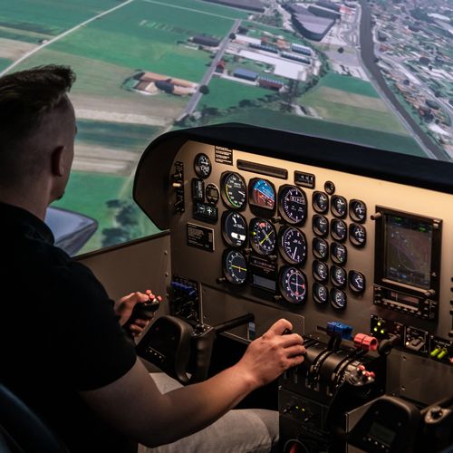 Pilot flying the Beechcraft Baron 58 simulator, hands on yoke and throttle, approach over green fields