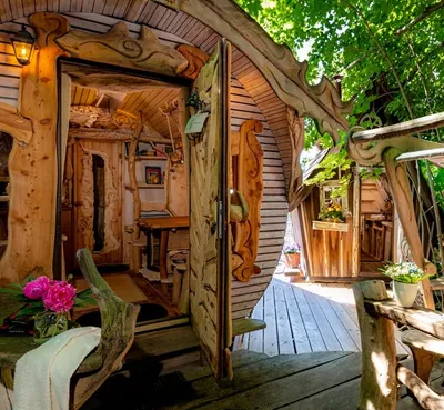 Open door of a barrel-shaped treehouse with carved wood details and flowers on the wooden deck