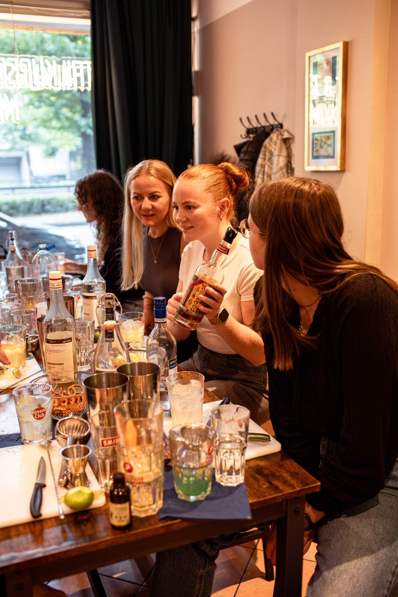 Group of four women gathered around a bar table filled with bottles and shakers, smiling together during a cocktail making course.