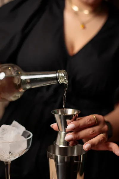 Close-up of hands pouring a clear spirit from a bottle into a jigger held over a cocktail shaker.