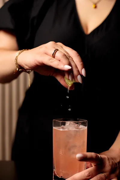 Close-up of a hand squeezing a lime wedge over a pink cocktail in a highball glass.