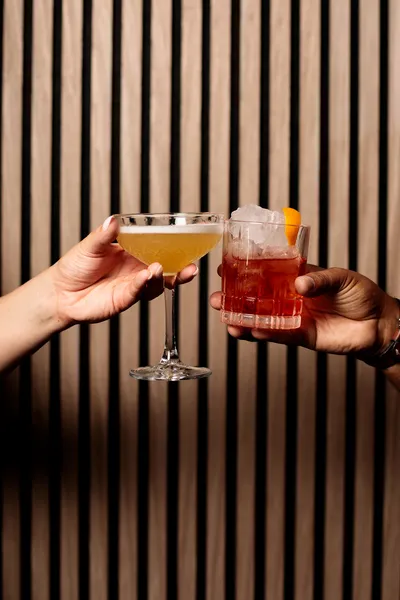 Two hands clinking together a yellow sour cocktail in a coupe glass and a red cocktail over ice garnished with orange peel.