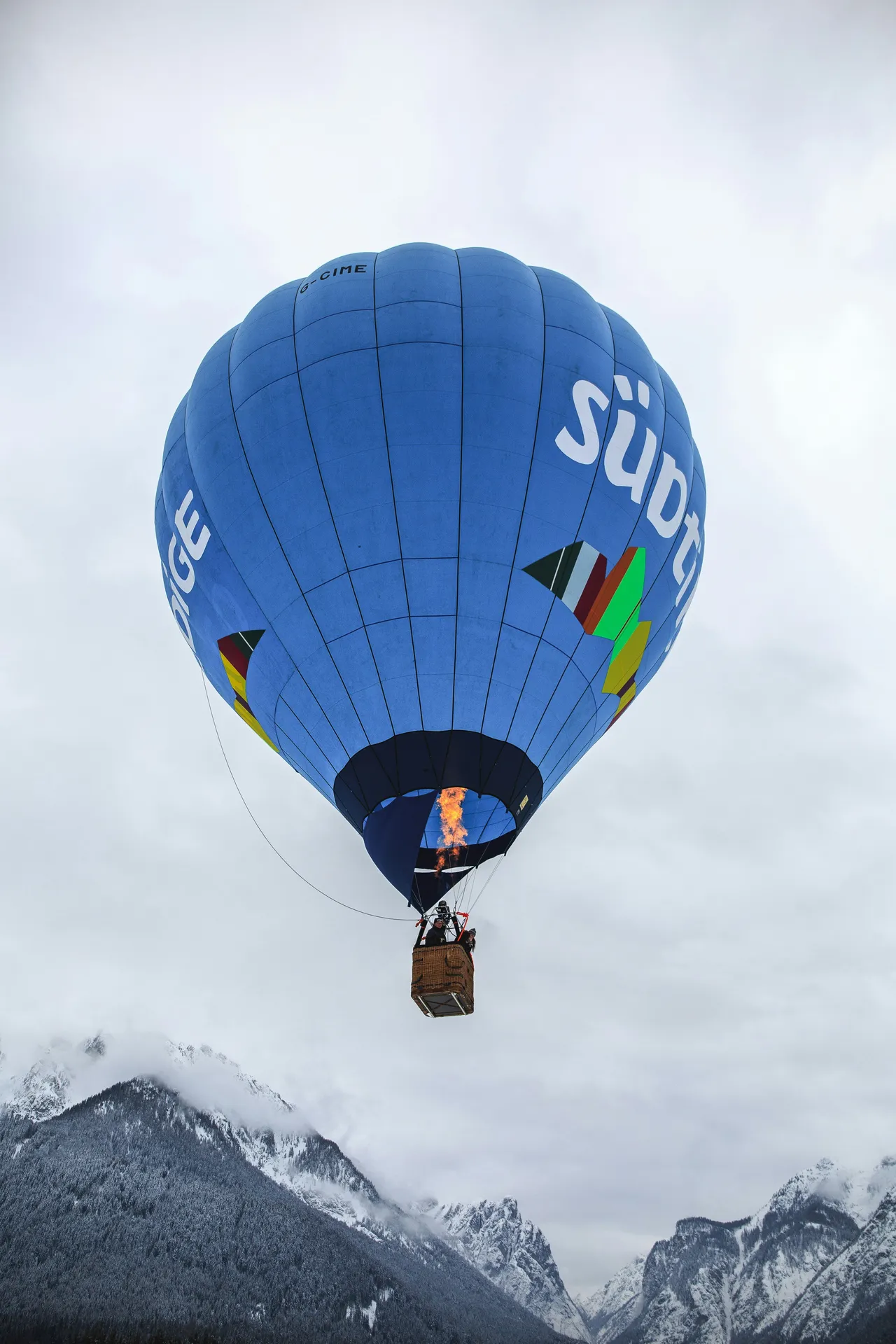 A blue hot air balloon with a lit burner flying above snow-covered mountains under an overcast sky.
