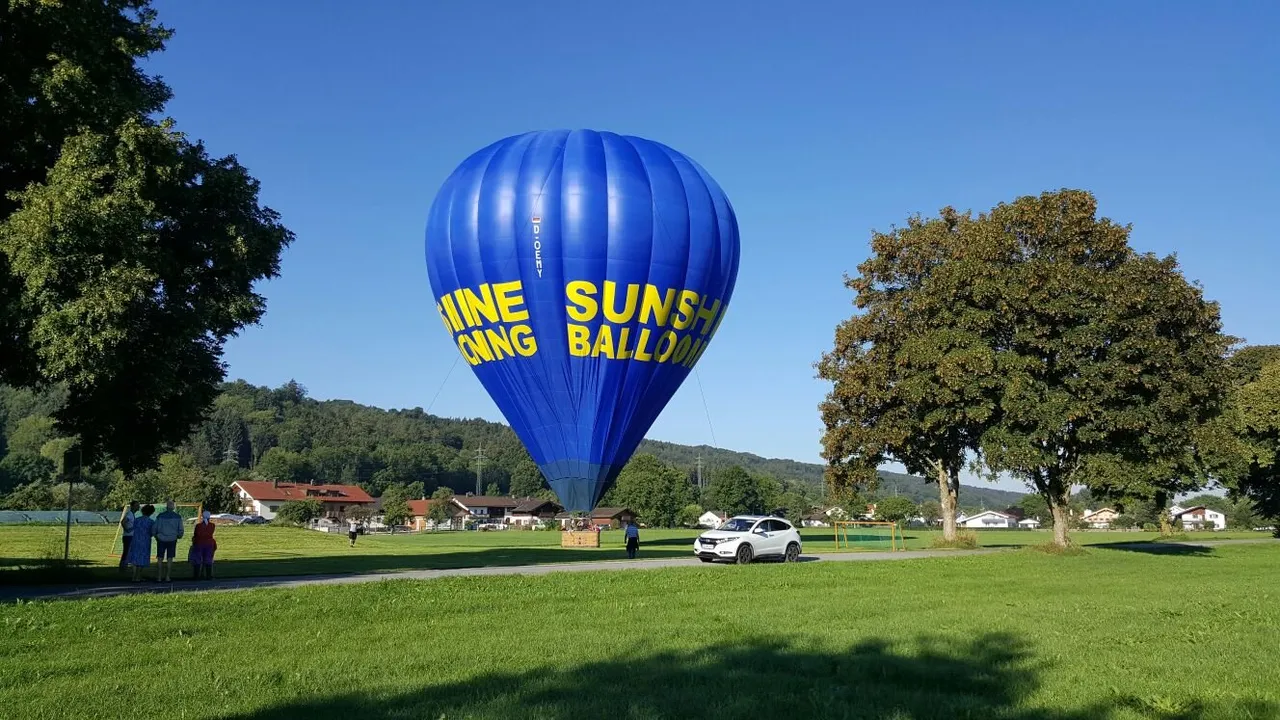 A bright blue hot air balloon with yellow text parked on a grassy field surrounded by trees and houses on a clear day.