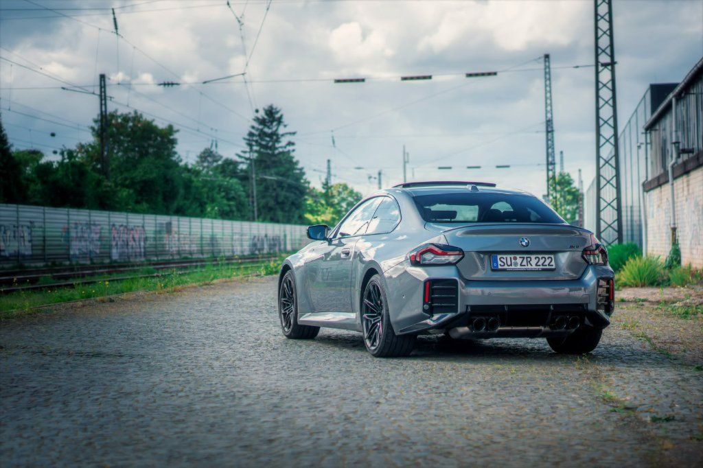 Grey BMW M2 Facelift 2026 seen from behind on a cobbled road beside railway tracks