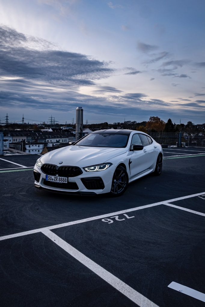 White BMW M8 Competition xDrive parked on a rooftop car park at dusk, seen from the front three-quarter angle