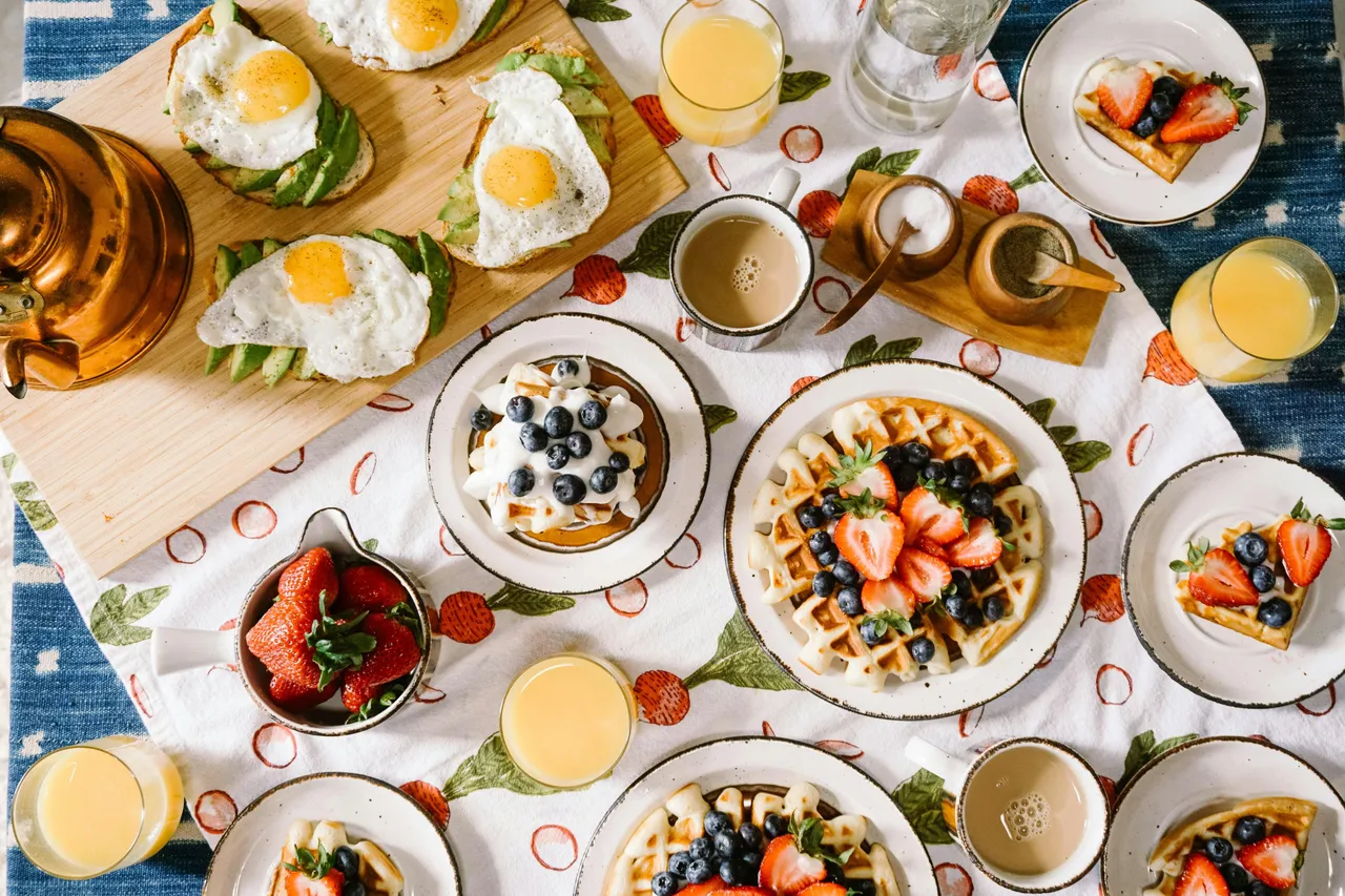 Beautiful brunch spread featuring waffles topped with strawberries and blueberries, fried eggs, coffee, fresh fruit, and orange juice arranged on a table.
