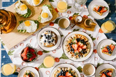 Beautiful brunch spread featuring waffles topped with strawberries and blueberries, fried eggs, coffee, fresh fruit, and orange juice arranged on a table.