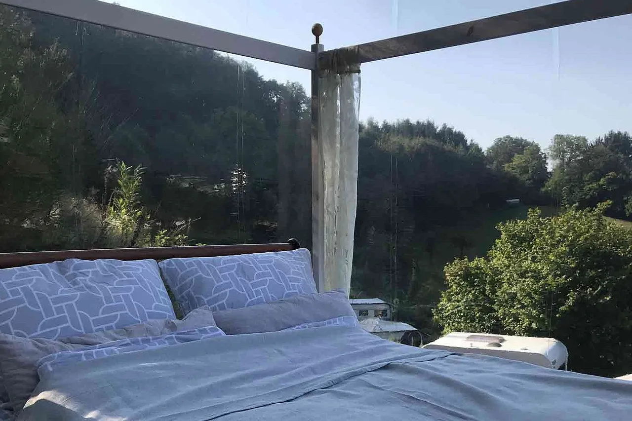 Bed with patterned pillows inside a transparent tent looking out onto green wooded hills