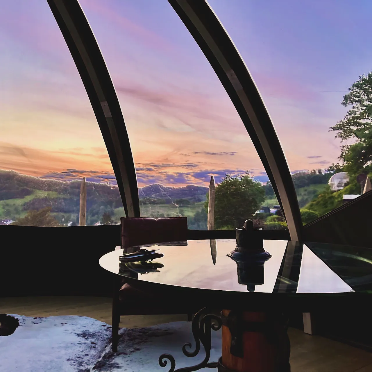 Inside a bubble tent with a glass table and leather chair, looking out over the hills at sunset