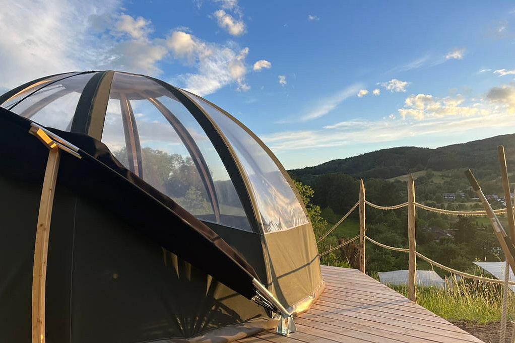 Partly transparent bubble tent on a wooden deck with rope railing overlooking wooded hills