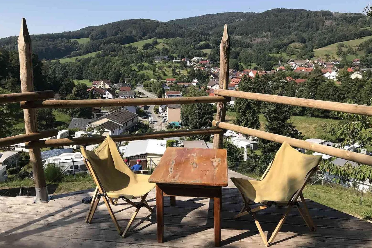 Wooden terrace with a table and two folding chairs overlooking a village and green hills