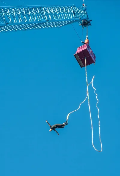 A person mid-dive during a bungee jump, suspended by a cord attached to a high crane platform against a clear blue sky.
