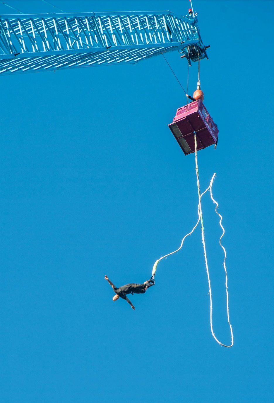 A person mid-dive during a bungee jump, suspended by a cord attached to a high crane platform against a clear blue sky.