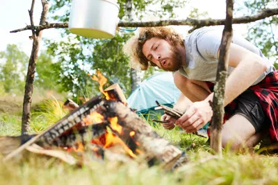 A man with curly hair kneels down to blow on a campfire under a cooking pot at a campsite.