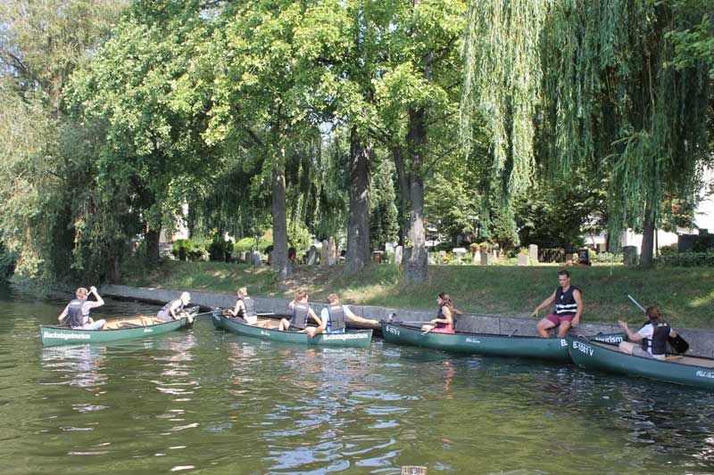 Several canoes with paddlers gliding along a willow-lined riverbank in Berlin