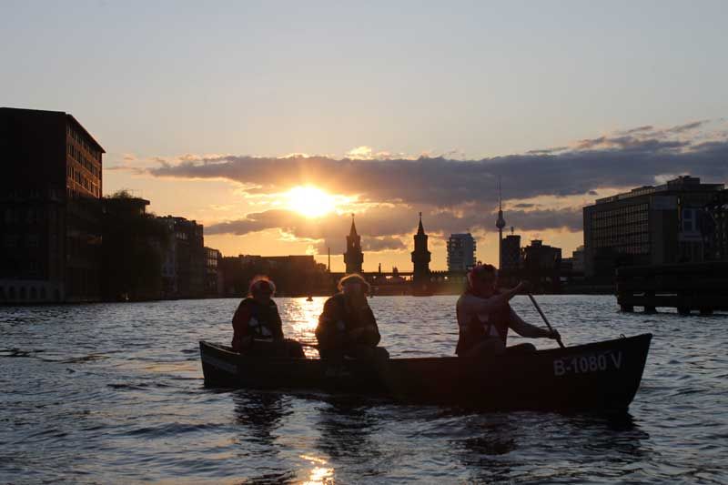 Three paddlers in a canoe silhouetted against the sunset near Oberbaum Bridge in Berlin