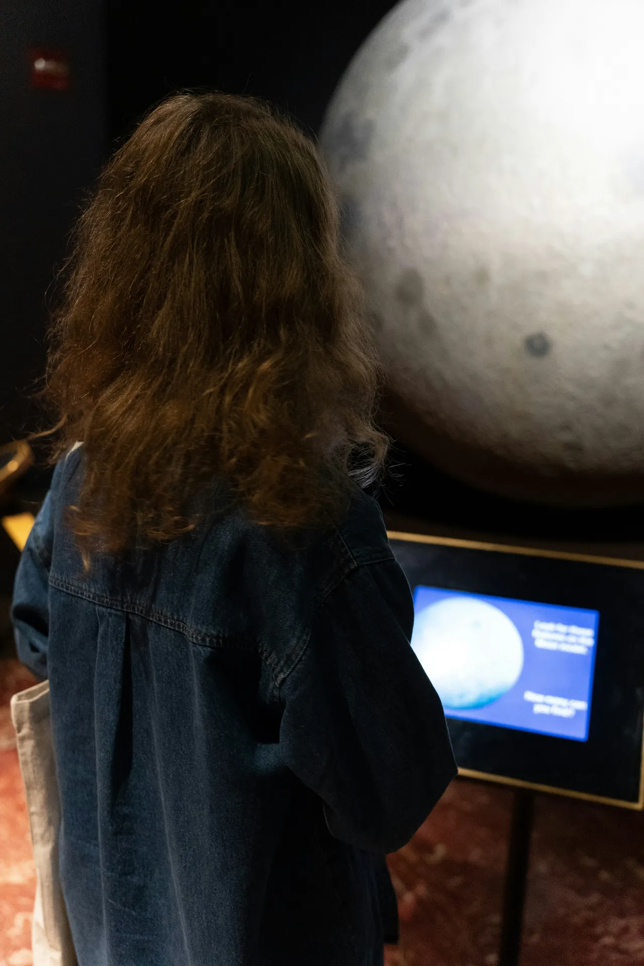 Rear view of a child with long brown hair wearing a denim jacket, looking at a large physical model of the Moon next to an interactive information screen at a museum exhibit.