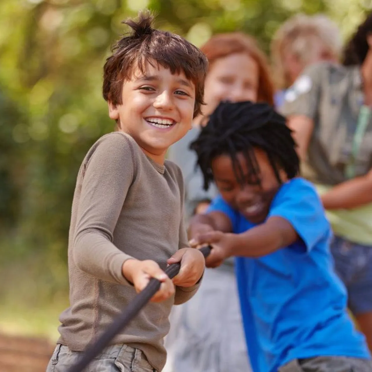 A cheerful young boy smiling at the camera while playing a game of tug-of-war with other children outdoors.