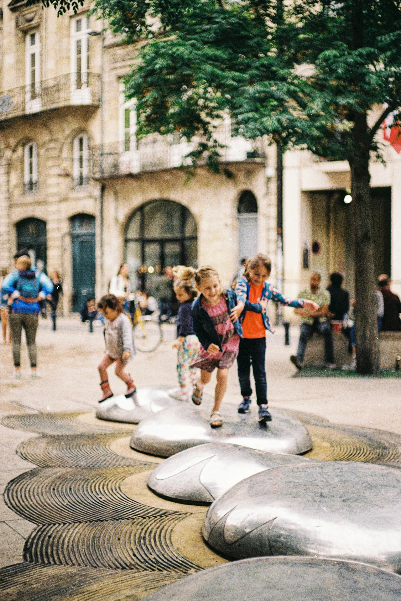 Young children playfully run and balance across smooth, metallic stepping stones embedded in a city plaza sidewalk, with historic European-style buildings and pedestrians in the background.