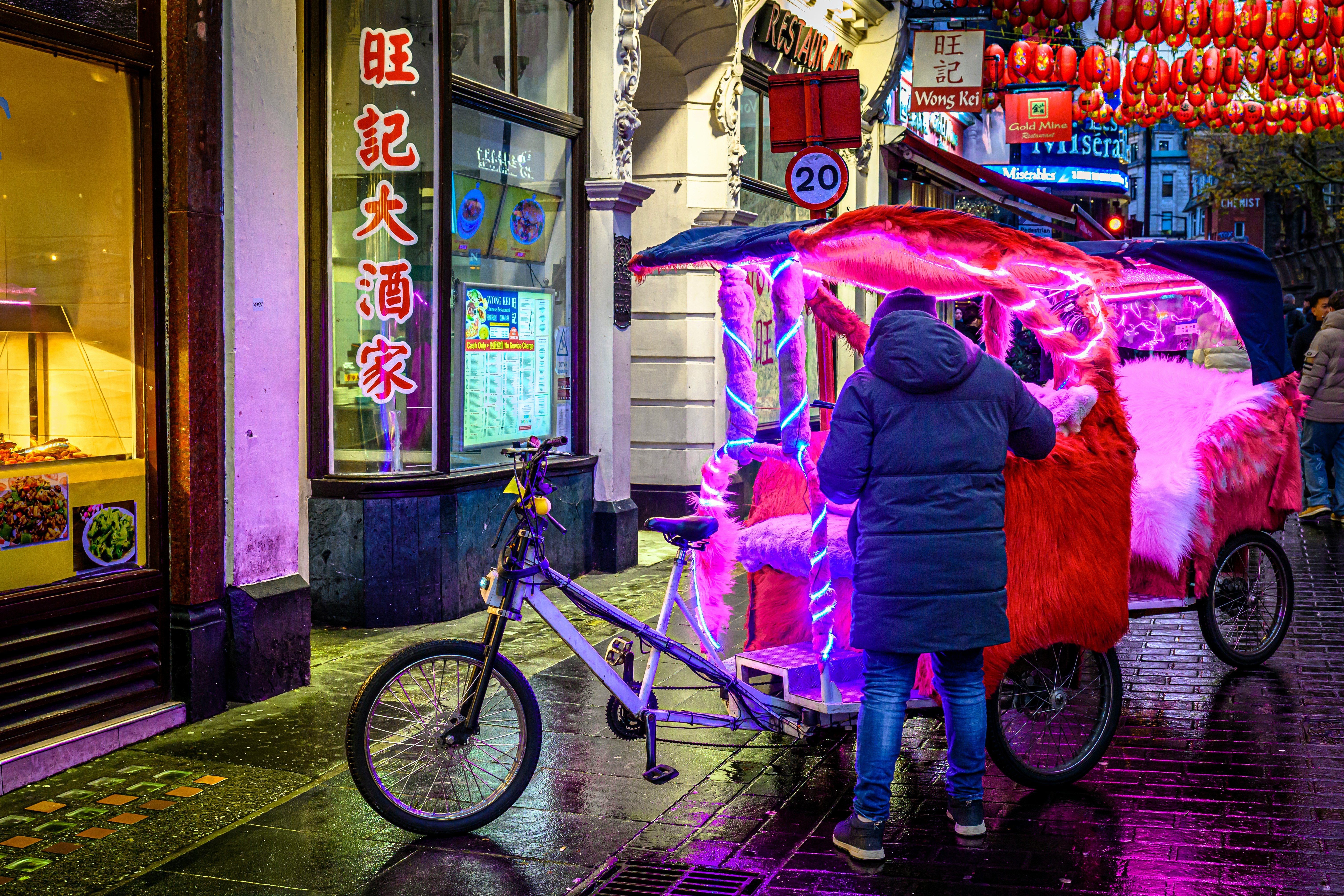 A person stands next to a vibrant, brightly illuminated pink cycle rickshaw decorated with faux fur and neon lights on a rainy street in Chinatown, next to a restaurant with Chinese signage.