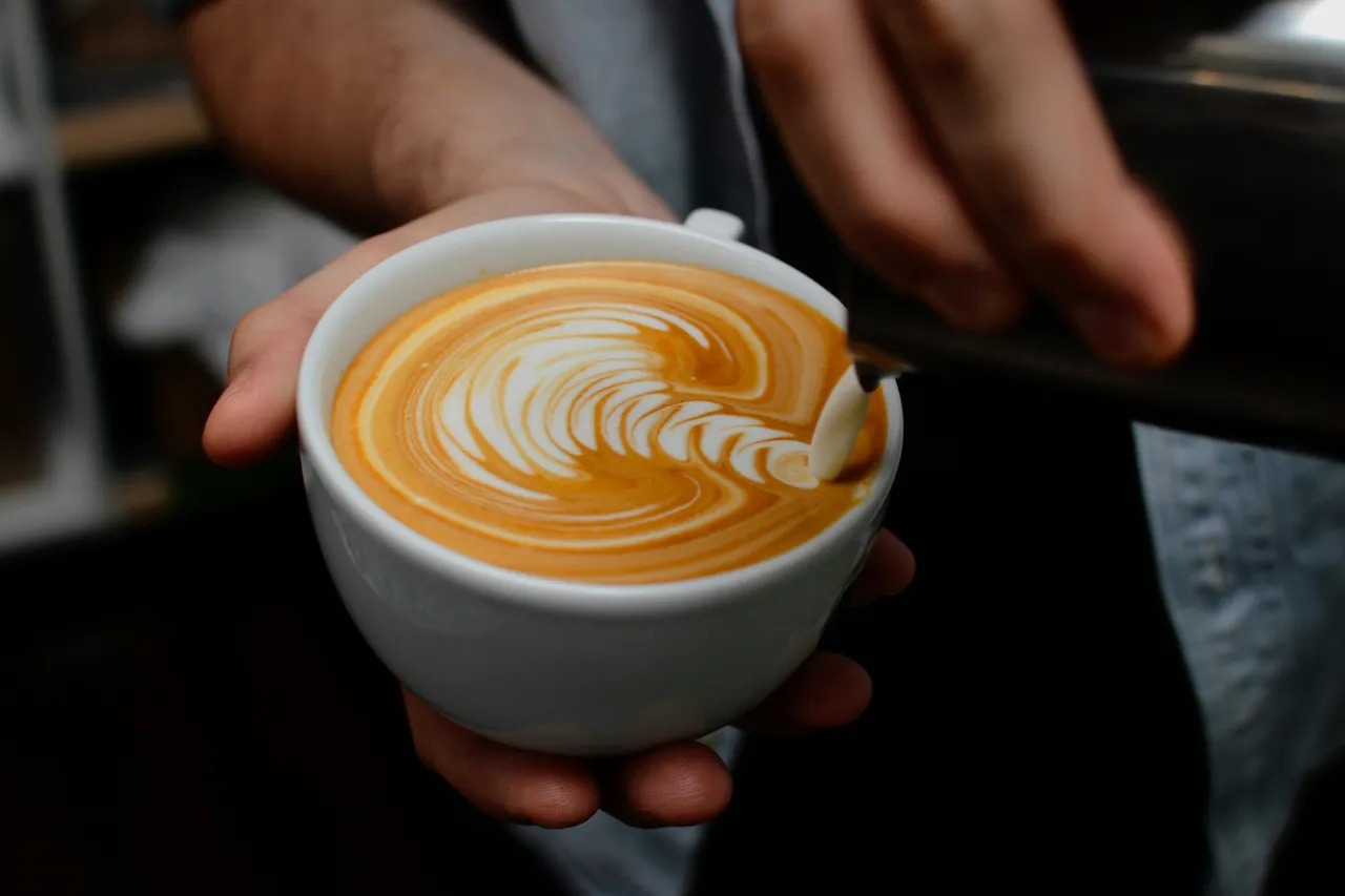 Hands holding a white ceramic cup of coffee with brown-and-white rosetta (rosettta leaf) latte art pattern in the crema.
