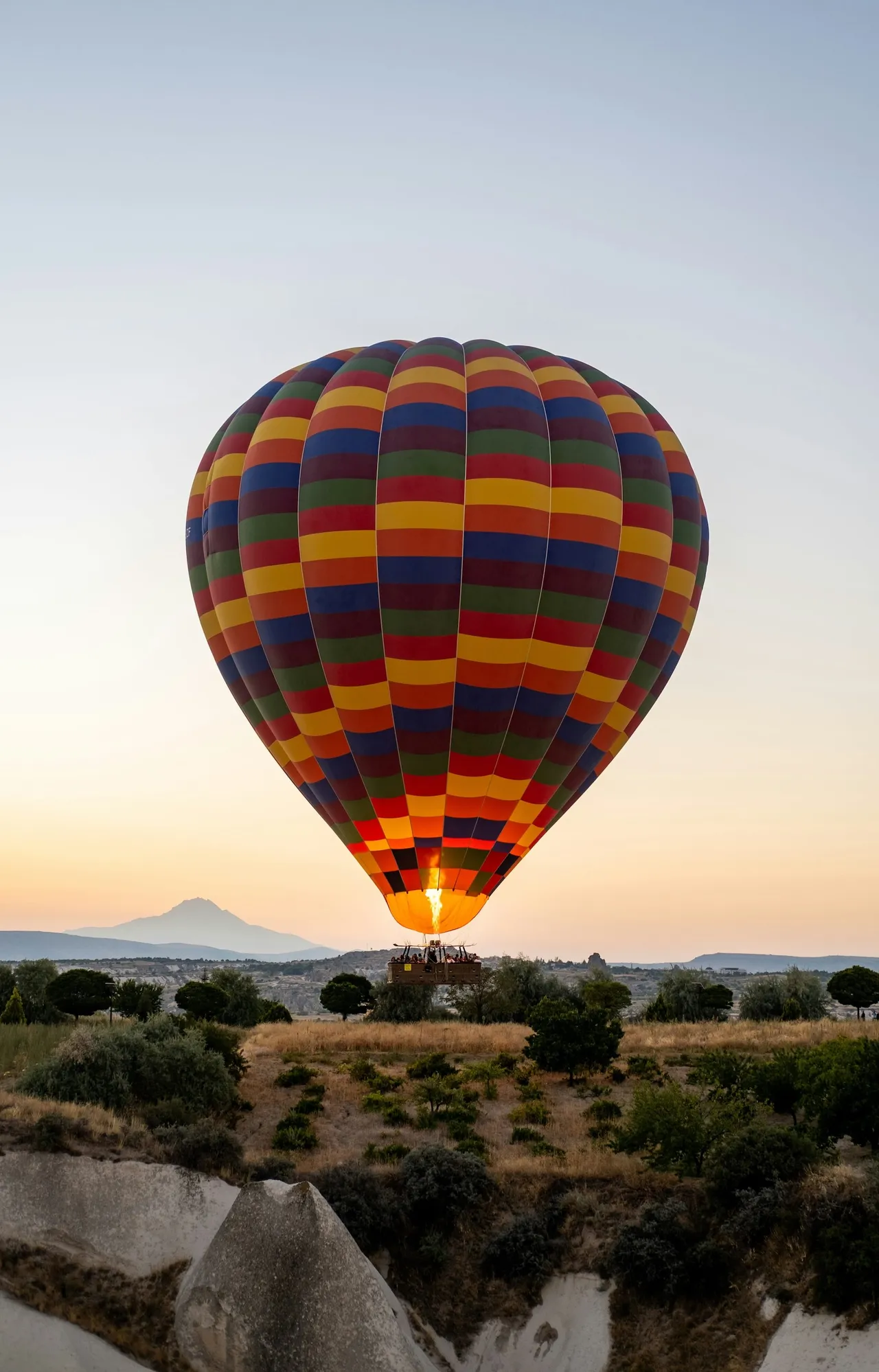 A colorful, multi-patterned hot air balloon flying low over a rocky landscape with trees and a distant mountain at sunrise.