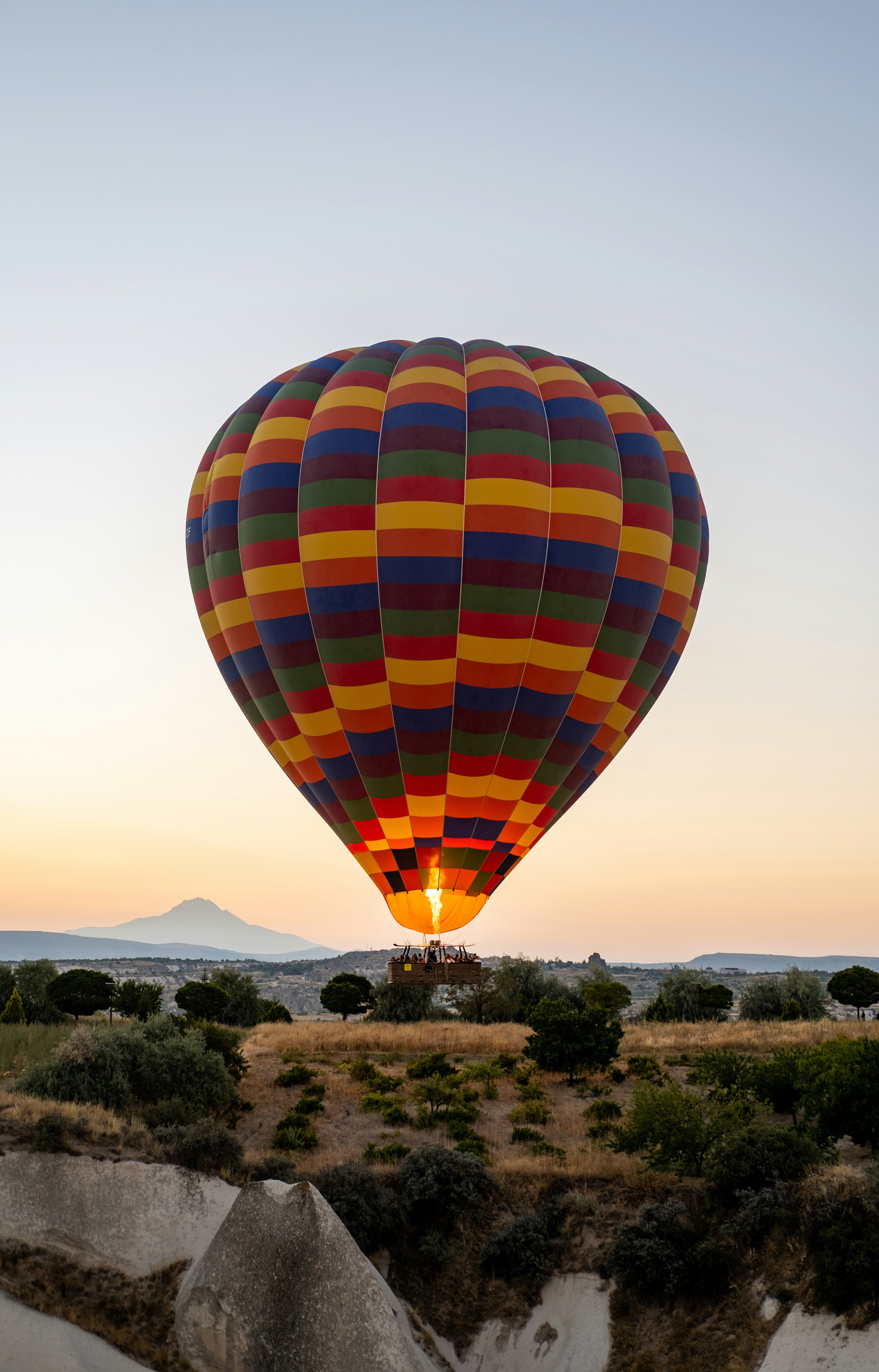 A colorful, multi-patterned hot air balloon flying low over a rocky landscape with trees and a distant mountain at sunrise.