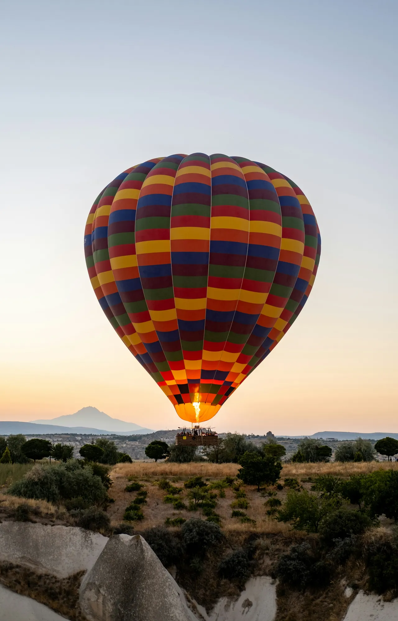 A colorful, multi-patterned hot air balloon flying low over a rocky landscape with trees and a distant mountain at sunrise.