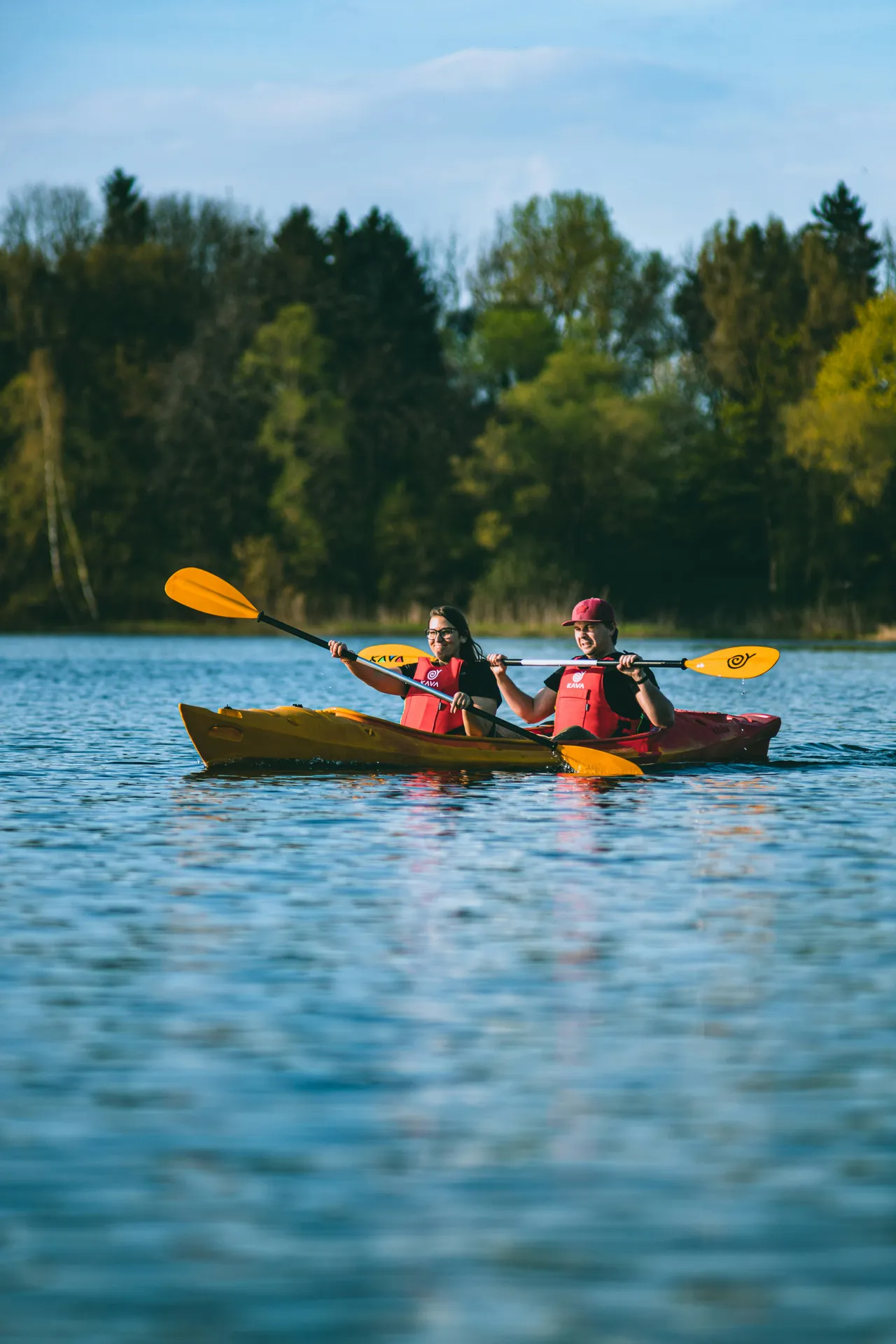 A man and a woman wearing red life jackets paddle together in a yellow tandem kayak on a calm lake with a forested shoreline.
