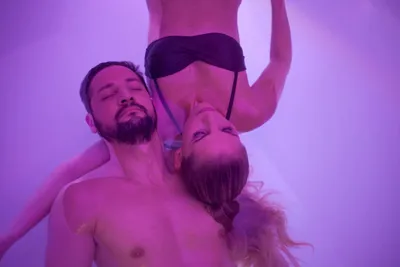 A man and woman with eyes closed in a close, relaxed pose under purple-toned lighting, evoking a calm spa or wellness atmosphere