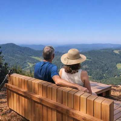 A man and a woman in a sun hat sitting on a wooden bench, looking out at a wide panorama of forested hills.