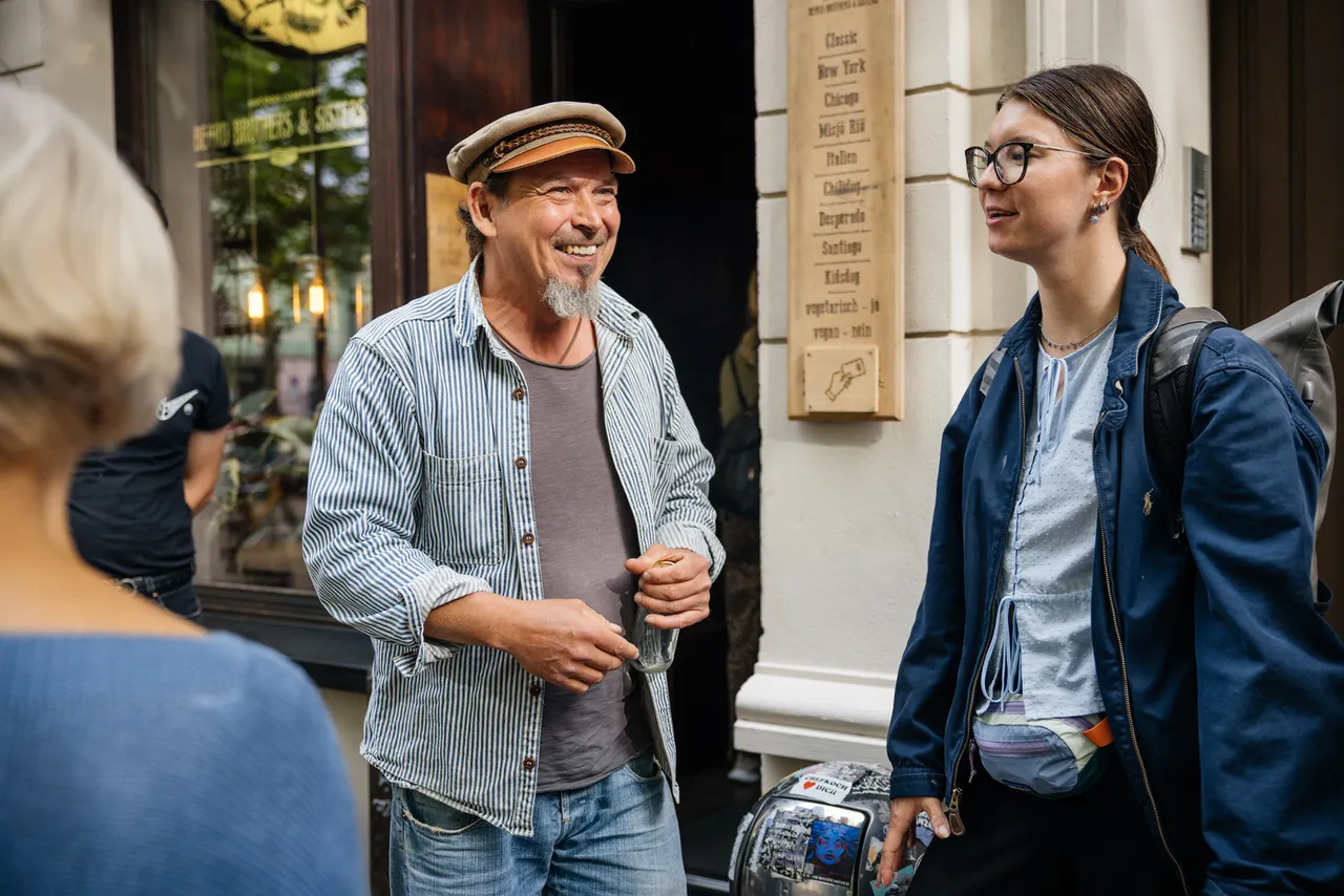 Smiling shop owner in a flat cap chatting with a tour guest outside his shop