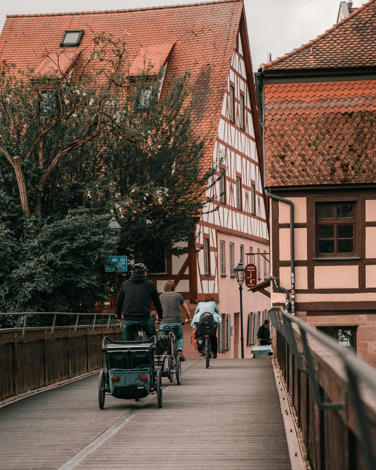Three cyclists ride away across a wooden bridge toward traditional timber-framed houses with red-tiled roofs, with one cyclist towing a bike trailer.