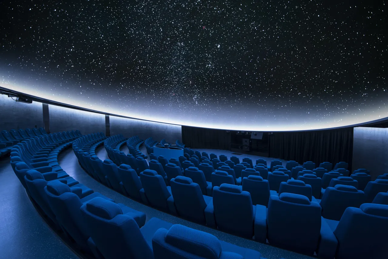 An empty planetarium theater with rows of curved blue seats facing a large domed ceiling displaying a realistic projection of a clear, starry night sky.