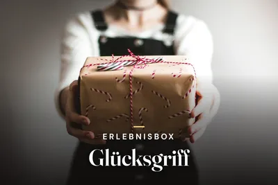 Person holding out a gift wrapped in brown paper with candy cane print and red ribbon, with the text "Erlebnisbox Glücksgriff"