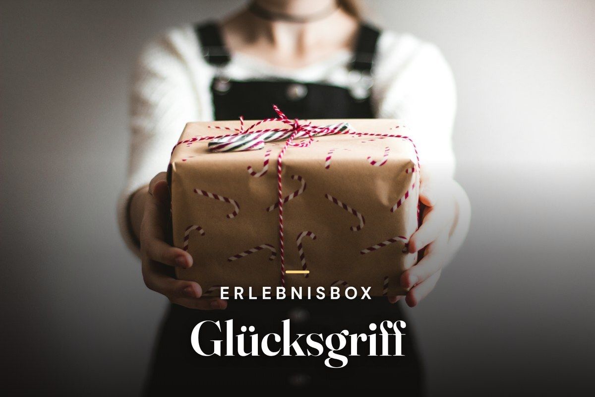 Person holding out a gift wrapped in brown paper with candy cane print and red ribbon, with the text "Erlebnisbox Glücksgriff"