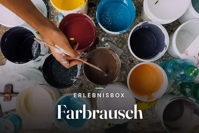 Hand dipping a brush into colorful paint buckets seen from above, with the text "Erlebnisbox Farbrausch"